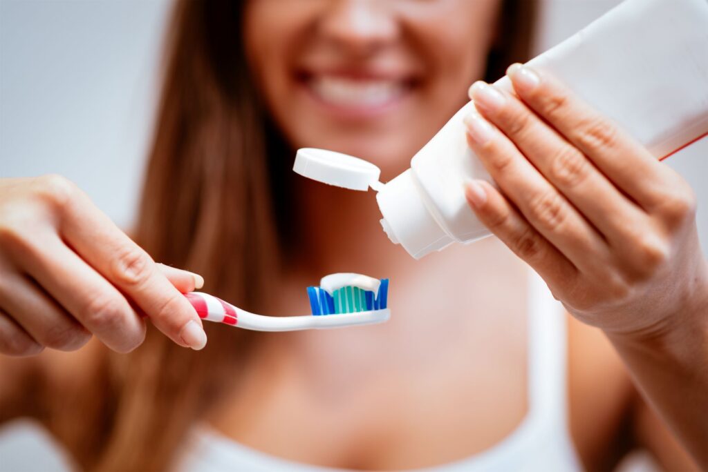 Woman blurry in background squeezing toothpaste onto toothbrush in foreground